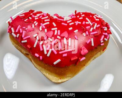 Un donut en forme de coeur rempli de pâte à gâteau, trempé dans le glaçage rouge, et recouvert de saupoudrés de Saint-Valentin festifs Banque D'Images