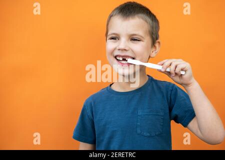 Un garçon sans la dent supérieure de lait dans un t-shirt bleu tient la brosse à dents à la main sur fond orange. Banque D'Images