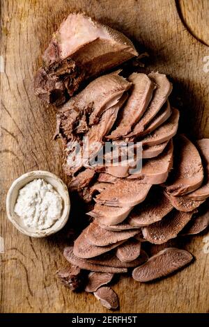 La langue de bœuf cuite au four est coupée en tranches et sert avec de la sauce au raifort sur une planche à découper en bois sur fond de bois. Pose à plat. Banque D'Images