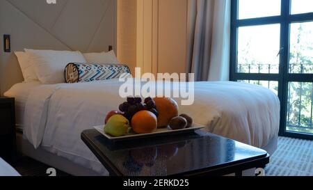 Une assiette de fruits sur une petite table à côté du confortable lit fraîchement préparé avec draps blancs, oreillers et literie Banque D'Images