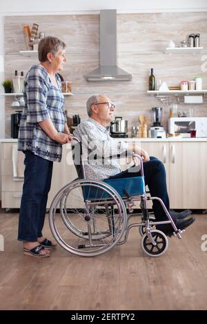 Femme poussant un homme de rêve invalide en fauteuil roulant dans la cuisine, discutant et souriant, appréciant la belle matinée ensemble. Heureux mature retraité paralysé handicapée homme positif Banque D'Images