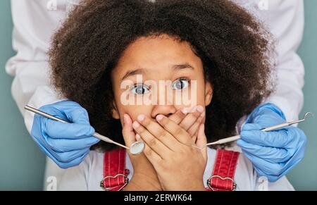 Petite fille afro-américaine effrayée à la rendez-vous du dentiste et couvre sa bouche avec les mains tout en examinant les dents Banque D'Images