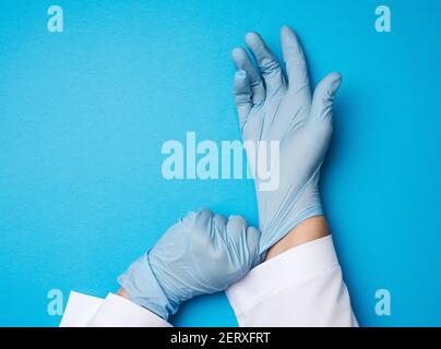 femme médecin dans un manteau blanc met sur ses mains des gants médicaux stériles en latex bleu sur ses mains, fond bleu, gros plan Banque D'Images