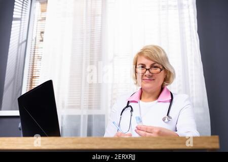 Portrait d'une femme médecin dans une robe médicale et des lunettes assis à la table, il utilise un ordinateur portable tout en consultant un patient. Banque D'Images