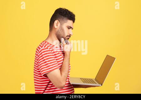 Portrait de profil pensif homme avec barbe en t-shirt rayé tenant un ordinateur portable et le menton frottant regardant l'affichage, pensant à de nouvelles affaires p Banque D'Images