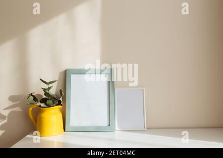 Un pot ou vase en céramique jaune avec des branches d'eucalyptus, des cadres photo blancs vides sur la table blanche à l'intérieur avec des murs beige près de la fenêtre. Banque D'Images