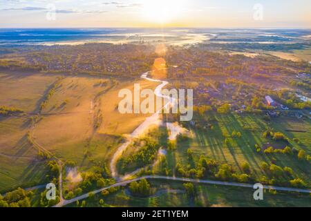 Survolant une vallée de rivière brumeuse, des prairies humides et de grands arbres qui jettent une ombre du soleil du matin. Vol au-dessus d'une petite ville rurale Banque D'Images