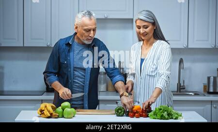 un couple interracial souriant prépare le petit-déjeuner avec des fruits et des légumes frais dans la cuisine Banque D'Images