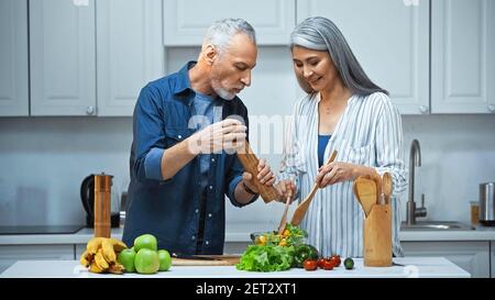 homme senior tenant un moulin à épices près de la bonne femme asiatique se préparant salade pour le petit déjeuner Banque D'Images