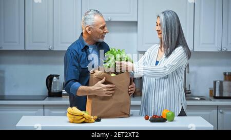 couple interracial âgé déballage sac en papier avec légumes frais et fruits dans la cuisine Banque D'Images