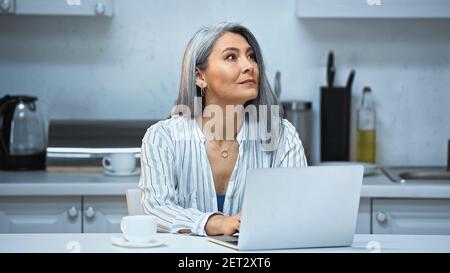 travailleur indépendant asiatique attentionné travaillant sur un ordinateur portable près d'une tasse de café dans la cuisine Banque D'Images