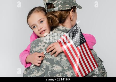 Fille souriante embrassant la mère en camouflage et tenant le drapeau américain isolé sur gris Banque D'Images