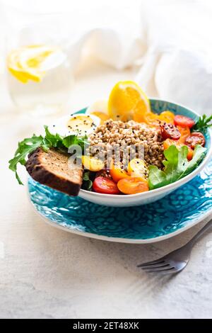 Bol de salade de sarrasin avec roquette, tomate, laitue, œuf dur et une tranche de pain à côté d'un verre d'eau de citron Banque D'Images