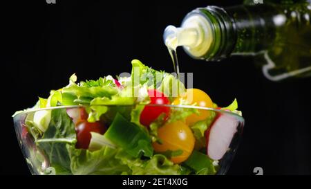 Huile d'olive versé de la bouteille sur la salade de légumes sur le noir arrière-plan Banque D'Images