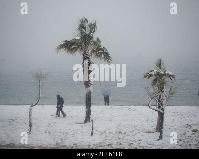 Deux personnes marchant et un homme seul avec un parapluie bleu debout près de la côte pendant une tempête de neige, près d'Athènes, Grèce, en février Banque D'Images