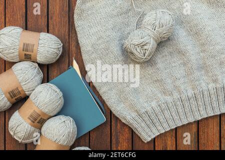 Vue de dessus des fils tricotés gris, carnet, crayon et une partie de pull tricoté sur fond de bois brun. Passe-temps et bricolage à la maison. Banque D'Images