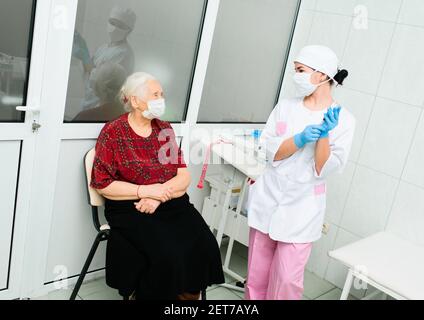 l'infirmière dans un masque médical met des gants en caoutchouc et se prépare à un examen médical d'un patient - an femme âgée à cheveux gris Banque D'Images