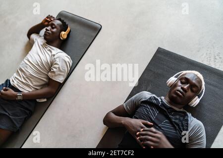 Deux jeunes hommes athlétiques afro-américains dans des écouteurs couchés tapis d'exercice après l'entraînement à l'intérieur Banque D'Images