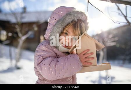 Petite fille à l'extérieur dans le jardin d'hiver, debout près d'un mangeoire à oiseaux en bois. Banque D'Images