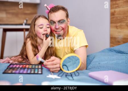 père et fille se maquillant ensemble, petite fille utilisant des cosmétiques décoratifs, s'amusant à jouer en appréciant le temps avec les parents à la maison le week-end Banque D'Images