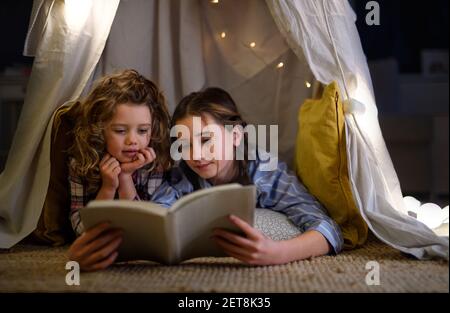 Sœurs à l'intérieur à la maison, en utilisant le livre de lecture en pyjama. Concept de verrouillage. Banque D'Images