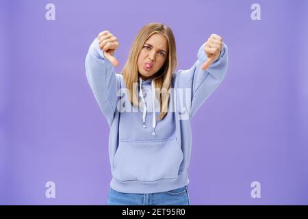 Photo en intérieur de moodie déplaisée et non impressionnée cool femme moderne avec les cheveux blonds et la peau bronzée tête inclinable en dehors la langue montre les pouces Banque D'Images