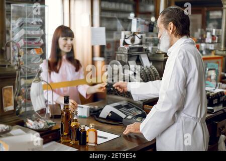 Vue latérale de l'homme caucasien sympathique barbu pharmacien offrant des médicaments à la jeune femme patient, payant pour le médicament avec carte de crédit. Sélectif Banque D'Images