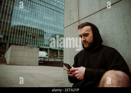 Athlète caucasien de sexe masculin dactylographiant sur un téléphone portable assis contre le mur après longue course Banque D'Images
