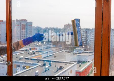 Une jeune femme méconnaissable en gants bleus lave le balcon fenêtre de l'extérieur avec un racleur spécial à long manche Banque D'Images