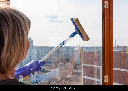 Une jeune femme méconnaissable en gants bleus lave la fenêtre de l'extérieur avec un racleur spécial sur un long poignée Banque D'Images
