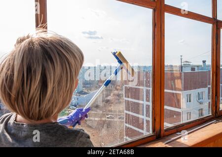 Une jeune femme méconnaissable en gants bleus lave le balcon fenêtre de l'extérieur avec un racleur spécial à long manche Banque D'Images