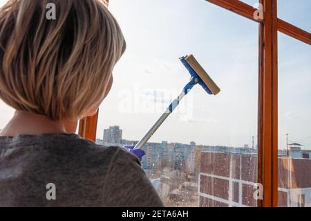Une jeune femme méconnaissable en gants bleus lave le balcon fenêtre de l'extérieur avec un racleur spécial à long manche Banque D'Images