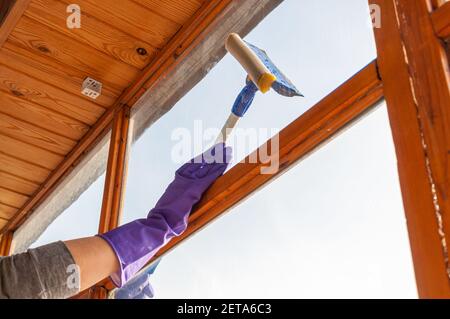 Une jeune femme méconnaissable en gants bleus lave le balcon fenêtre de l'extérieur avec un racleur spécial à long manche Banque D'Images