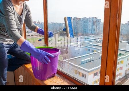 Une jeune femme méconnaissable en gants bleus lave le balcon fenêtre de l'extérieur avec un racleur spécial à long manche Banque D'Images