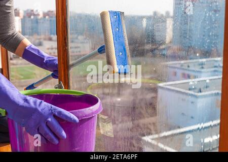 Une jeune femme méconnaissable en gants bleus lave le balcon fenêtre de l'extérieur avec un racleur spécial à long manche Banque D'Images
