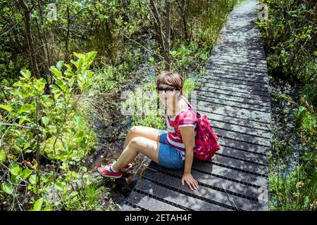 Une femme avec sac à dos assise sur le chemin en bois et se détendre après la randonnée dans la forêt d'été. Apprécier, unité avec la nature, détox numérique Banque D'Images