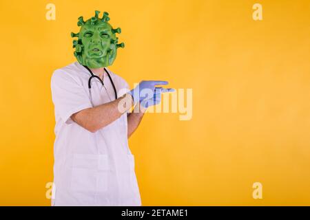Médecin - infirmière en coronavirus déguisée portant un masque en latex - virus Covid-19 pointant de côté sur fond jaune. Concept de coronavirus Banque D'Images