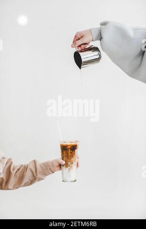 Main de la femme versant le café du pot métallique dans le verre avec du lait pour préparer des boissons au cappuccino latte pour le petit déjeuner sur fond de mur léger. Concept de boissons au café Banque D'Images