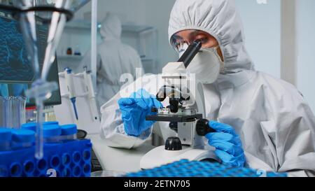 Une chercheuse porte une combinaison d'epi et regarde des échantillons au microscope dans un laboratoire équipé. Médecin travaillant avec diverses bactéries, tissus, analyses de sang, recherche pharmaceutique pour les antibiotiques Banque D'Images
