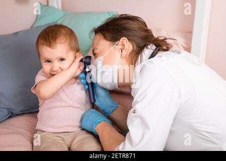 Le pédiatre examine l'oreille de la petite fille à la maison pendant la quarantaine pandémique COVID-19 du coronavirus. Médecin utilisant l'otoscope (auriscope) pour vérifier le canal auditif a Banque D'Images
