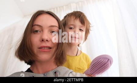 Mignon petit garçon brosses bonne mère cheveux courts regarder les uns les autres pendant la vidéo près de la fenêtre dans la pièce lumineuse de près Banque D'Images