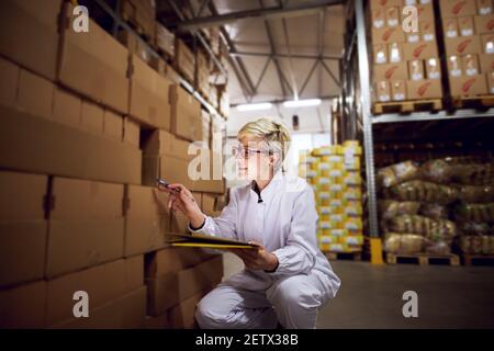 Jeune femme joyeuse et heureuse dans la zone de stockage de l'installation vérifie les boîtes dans la zone de stockage tout en s'agenouillant et en tenant une chemise jaune. Banque D'Images