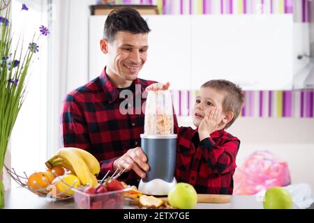 Le jeune père et le fils heureux mélangent des fruits frais divers sur le comptoir de cuisine tout en portant la même chemise rouge. Banque D'Images