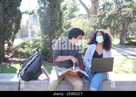 Deux étudiants latins portant un masque de protection étudiant ensemble assis sur un banc à l'extérieur. Nouvelle norme sur le campus universitaire. Banque D'Images