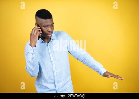 Jeune homme afro-américain portant une chemise décontractée fond jaune parlant sur le téléphone avec une expression inquiète Banque D'Images