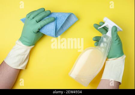 Les mains en gants verts maintiennent le chiffon et les détergents sur fond jaune. Concept de nettoyage ou d'entretien ménager. Pose à plat. Vue de dessus Banque D'Images