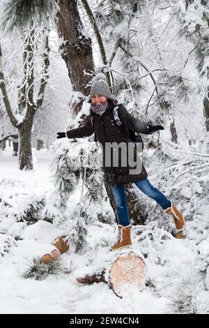 Jeune femme souriante debout sur un arbre déchu après avoir traîneaux et neige dans un parc d'hiver enneigé. Fille appréciant l'hiver enneigé, jour gelé. Entrez Banque D'Images