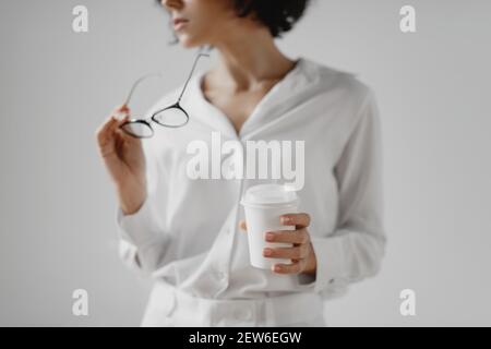 Image rognée d'une jeune femme tenant des lunettes et buvant du café dans une tasse de papier sur fond blanc. Banque D'Images