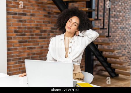 Femme indépendante élégante de course mixte avec coiffure afro assis au bureau avec les yeux fermés dans le bureau moderne, se sentant fatiguée du travail constant sur ordinateur portable, se reposant, étirant le cou avec la main Banque D'Images