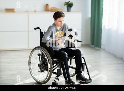 Un joueur de football adolescent handicapé avec balle et trophée assis en fauteuil roulant, se sentant déprimé au-dessus de sa blessure à la maison Banque D'Images